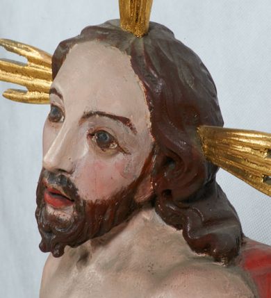 Zdjęcie nr 4: Rzeźba Chrystusa Zmartwychwstałego pełnoplastyczna, wolnostojąca, ustawiona na niskim cokole. Chrystus ukazany w całej postaci, ustawiony frontalnie, w delikatnym kontrapoście, ma prawą nogę ugiętą w kolanie i wspartą na czaszce z wężem. Ręce ma ugięte w łokciach i wyciągnięte przed siebie, w lewej trzyma chorągiew o długim drzewcu, a prawą unosi w geście błogosławieństwa. Jego twarz jest pociągła, o wysokim czole, długim i wąskim nosie, dużych oczach i rozchylonych ustach, okolona średniej długości, ciemną brodą i długimi, falowanymi włosami, które opadają na plecy i lewe ramię. Wokół głowy ma złoty nimb krzyżowy utworzony z wiązek promieni o nierównej długości. Chrystus ubrany jest w białe perizonium oraz czerwony płaszcz odsłaniający tors, otaczający figurę i przerzucony przez lewe ramię. Na dłoniach, stopach i boku zaznaczone są ślady męki. Polichromia ciała jest naturalistyczna, nimb pozłacany, drzewce chorągwi srebrzone.

