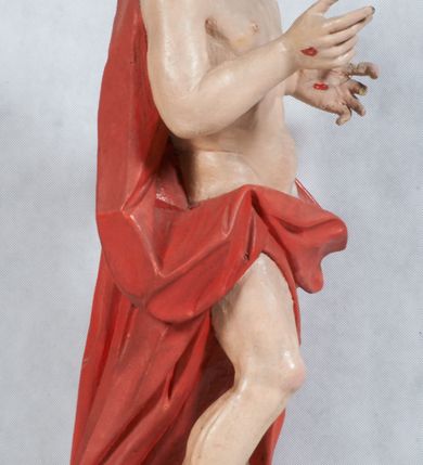 Zdjęcie nr 10: Rzeźba Chrystusa Zmartwychwstałego pełnoplastyczna, wolnostojąca, ustawiona na niskim cokole. Chrystus ukazany w całej postaci, ustawiony frontalnie, w delikatnym kontrapoście, ma prawą nogę ugiętą w kolanie i wspartą na czaszce z wężem. Ręce ma ugięte w łokciach i wyciągnięte przed siebie, w lewej trzyma chorągiew o długim drzewcu, a prawą unosi w geście błogosławieństwa. Jego twarz jest pociągła, o wysokim czole, długim i wąskim nosie, dużych oczach i rozchylonych ustach, okolona średniej długości, ciemną brodą i długimi, falowanymi włosami, które opadają na plecy i lewe ramię. Wokół głowy ma złoty nimb krzyżowy utworzony z wiązek promieni o nierównej długości. Chrystus ubrany jest w białe perizonium oraz czerwony płaszcz odsłaniający tors, otaczający figurę i przerzucony przez lewe ramię. Na dłoniach, stopach i boku zaznaczone są ślady męki. Polichromia ciała jest naturalistyczna, nimb pozłacany, drzewce chorągwi srebrzone.

