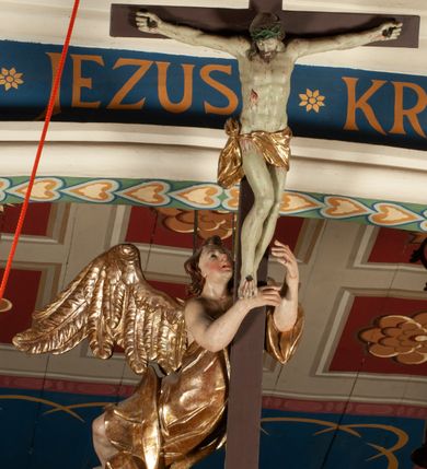 Zdjęcie nr 1: Krucyfiks zawieszony w łuku tęczowym kościoła. Do prostego krzyża przybita trzema gwoździami figura umarłego Chrystusa. Postać ukazana z szeroko i prosto rozłożonymi ramionami, z głową opadającą delikatnie w dół i z prawą nogą ugiętą w kolanie. Twarz Chrystusa jest podłużna, o zamkniętych oczach, długim i wąskim nosie, okolona jest krótką brodą rozdzieloną na końcu na dwa małe pukle i długimi włosami opadającymi na plecy i prawe ramię. Na głowie szeroka, zielona korona cierniowa. Ciało o delikatnie podkreślonej anatomii, smukłe. Perizonium zawiązane na prawym boku, odsłaniające prawe udo, z jednym końcem zwisającym wzdłuż prawego boku. Krzyż prosty, gładki, malowany na kolor ciemnobrązowy. Po jego lewej stronie, od tyłu nadlatujący anioł, nadnaturalnej wielkości, obejmujący rękami krzyż. Anioł ubrany w długą, złotą suknię, z rozłożonymi skrzydłami. Twarz anioła owalna, z otwartymi ustami, wzrokiem skierowanym na Chrystusa, nosem długim i spiczastym, silnie zarumienionymi policzkami, kręconymi włosami średniej długości. Nad głową Chrystusa na pionowej belce złota banderola z napisem „I.N./R.I.” Polichromia w odsłoniętych partiach ciała naturalistyczna, na figurze Chrystusa w odcieniach szarości, szaty i detale złocone. 
