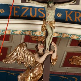 Zdjęcie nr 1: Krucyfiks zawieszony w łuku tęczowym kościoła. Do prostego krzyża przybita trzema gwoździami figura umarłego Chrystusa. Postać ukazana z szeroko i prosto rozłożonymi ramionami, z głową opadającą delikatnie w dół i z prawą nogą ugiętą w kolanie. Twarz Chrystusa jest podłużna, o zamkniętych oczach, długim i wąskim nosie, okolona jest krótką brodą rozdzieloną na końcu na dwa małe pukle i długimi włosami opadającymi na plecy i prawe ramię. Na głowie szeroka, zielona korona cierniowa. Ciało o delikatnie podkreślonej anatomii, smukłe. Perizonium zawiązane na prawym boku, odsłaniające prawe udo, z jednym końcem zwisającym wzdłuż prawego boku. Krzyż prosty, gładki, malowany na kolor ciemnobrązowy. Po jego lewej stronie, od tyłu nadlatujący anioł, nadnaturalnej wielkości, obejmujący rękami krzyż. Anioł ubrany w długą, złotą suknię, z rozłożonymi skrzydłami. Twarz anioła owalna, z otwartymi ustami, wzrokiem skierowanym na Chrystusa, nosem długim i spiczastym, silnie zarumienionymi policzkami, kręconymi włosami średniej długości. Nad głową Chrystusa na pionowej belce złota banderola z napisem „I.N./R.I.” Polichromia w odsłoniętych partiach ciała naturalistyczna, na figurze Chrystusa w odcieniach szarości, szaty i detale złocone. 
