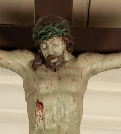 Zdjęcie nr 17: Krucyfiks zawieszony w łuku tęczowym kościoła. Do prostego krzyża przybita trzema gwoździami figura umarłego Chrystusa. Postać ukazana z szeroko i prosto rozłożonymi ramionami, z głową opadającą delikatnie w dół i z prawą nogą ugiętą w kolanie. Twarz Chrystusa jest podłużna, o zamkniętych oczach, długim i wąskim nosie, okolona jest krótką brodą rozdzieloną na końcu na dwa małe pukle i długimi włosami opadającymi na plecy i prawe ramię. Na głowie szeroka, zielona korona cierniowa. Ciało o delikatnie podkreślonej anatomii, smukłe. Perizonium zawiązane na prawym boku, odsłaniające prawe udo, z jednym końcem zwisającym wzdłuż prawego boku. Krzyż prosty, gładki, malowany na kolor ciemnobrązowy. Po jego lewej stronie, od tyłu nadlatujący anioł, nadnaturalnej wielkości, obejmujący rękami krzyż. Anioł ubrany w długą, złotą suknię, z rozłożonymi skrzydłami. Twarz anioła owalna, z otwartymi ustami, wzrokiem skierowanym na Chrystusa, nosem długim i spiczastym, silnie zarumienionymi policzkami, kręconymi włosami średniej długości. Nad głową Chrystusa na pionowej belce złota banderola z napisem „I.N./R.I.” Polichromia w odsłoniętych partiach ciała naturalistyczna, na figurze Chrystusa w odcieniach szarości, szaty i detale złocone. 
