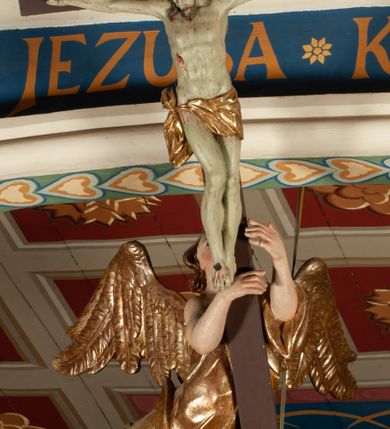 Zdjęcie nr 13: Krucyfiks zawieszony w łuku tęczowym kościoła. Do prostego krzyża przybita trzema gwoździami figura umarłego Chrystusa. Postać ukazana z szeroko i prosto rozłożonymi ramionami, z głową opadającą delikatnie w dół i z prawą nogą ugiętą w kolanie. Twarz Chrystusa jest podłużna, o zamkniętych oczach, długim i wąskim nosie, okolona jest krótką brodą rozdzieloną na końcu na dwa małe pukle i długimi włosami opadającymi na plecy i prawe ramię. Na głowie szeroka, zielona korona cierniowa. Ciało o delikatnie podkreślonej anatomii, smukłe. Perizonium zawiązane na prawym boku, odsłaniające prawe udo, z jednym końcem zwisającym wzdłuż prawego boku. Krzyż prosty, gładki, malowany na kolor ciemnobrązowy. Po jego lewej stronie, od tyłu nadlatujący anioł, nadnaturalnej wielkości, obejmujący rękami krzyż. Anioł ubrany w długą, złotą suknię, z rozłożonymi skrzydłami. Twarz anioła owalna, z otwartymi ustami, wzrokiem skierowanym na Chrystusa, nosem długim i spiczastym, silnie zarumienionymi policzkami, kręconymi włosami średniej długości. Nad głową Chrystusa na pionowej belce złota banderola z napisem „I.N./R.I.” Polichromia w odsłoniętych partiach ciała naturalistyczna, na figurze Chrystusa w odcieniach szarości, szaty i detale złocone. 
