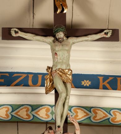 Zdjęcie nr 16: Krucyfiks zawieszony w łuku tęczowym kościoła. Do prostego krzyża przybita trzema gwoździami figura umarłego Chrystusa. Postać ukazana z szeroko i prosto rozłożonymi ramionami, z głową opadającą delikatnie w dół i z prawą nogą ugiętą w kolanie. Twarz Chrystusa jest podłużna, o zamkniętych oczach, długim i wąskim nosie, okolona jest krótką brodą rozdzieloną na końcu na dwa małe pukle i długimi włosami opadającymi na plecy i prawe ramię. Na głowie szeroka, zielona korona cierniowa. Ciało o delikatnie podkreślonej anatomii, smukłe. Perizonium zawiązane na prawym boku, odsłaniające prawe udo, z jednym końcem zwisającym wzdłuż prawego boku. Krzyż prosty, gładki, malowany na kolor ciemnobrązowy. Po jego lewej stronie, od tyłu nadlatujący anioł, nadnaturalnej wielkości, obejmujący rękami krzyż. Anioł ubrany w długą, złotą suknię, z rozłożonymi skrzydłami. Twarz anioła owalna, z otwartymi ustami, wzrokiem skierowanym na Chrystusa, nosem długim i spiczastym, silnie zarumienionymi policzkami, kręconymi włosami średniej długości. Nad głową Chrystusa na pionowej belce złota banderola z napisem „I.N./R.I.” Polichromia w odsłoniętych partiach ciała naturalistyczna, na figurze Chrystusa w odcieniach szarości, szaty i detale złocone. 
