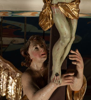 Zdjęcie nr 14: Krucyfiks zawieszony w łuku tęczowym kościoła. Do prostego krzyża przybita trzema gwoździami figura umarłego Chrystusa. Postać ukazana z szeroko i prosto rozłożonymi ramionami, z głową opadającą delikatnie w dół i z prawą nogą ugiętą w kolanie. Twarz Chrystusa jest podłużna, o zamkniętych oczach, długim i wąskim nosie, okolona jest krótką brodą rozdzieloną na końcu na dwa małe pukle i długimi włosami opadającymi na plecy i prawe ramię. Na głowie szeroka, zielona korona cierniowa. Ciało o delikatnie podkreślonej anatomii, smukłe. Perizonium zawiązane na prawym boku, odsłaniające prawe udo, z jednym końcem zwisającym wzdłuż prawego boku. Krzyż prosty, gładki, malowany na kolor ciemnobrązowy. Po jego lewej stronie, od tyłu nadlatujący anioł, nadnaturalnej wielkości, obejmujący rękami krzyż. Anioł ubrany w długą, złotą suknię, z rozłożonymi skrzydłami. Twarz anioła owalna, z otwartymi ustami, wzrokiem skierowanym na Chrystusa, nosem długim i spiczastym, silnie zarumienionymi policzkami, kręconymi włosami średniej długości. Nad głową Chrystusa na pionowej belce złota banderola z napisem „I.N./R.I.” Polichromia w odsłoniętych partiach ciała naturalistyczna, na figurze Chrystusa w odcieniach szarości, szaty i detale złocone. 
