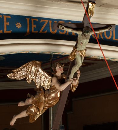 Zdjęcie nr 12: Krucyfiks zawieszony w łuku tęczowym kościoła. Do prostego krzyża przybita trzema gwoździami figura umarłego Chrystusa. Postać ukazana z szeroko i prosto rozłożonymi ramionami, z głową opadającą delikatnie w dół i z prawą nogą ugiętą w kolanie. Twarz Chrystusa jest podłużna, o zamkniętych oczach, długim i wąskim nosie, okolona jest krótką brodą rozdzieloną na końcu na dwa małe pukle i długimi włosami opadającymi na plecy i prawe ramię. Na głowie szeroka, zielona korona cierniowa. Ciało o delikatnie podkreślonej anatomii, smukłe. Perizonium zawiązane na prawym boku, odsłaniające prawe udo, z jednym końcem zwisającym wzdłuż prawego boku. Krzyż prosty, gładki, malowany na kolor ciemnobrązowy. Po jego lewej stronie, od tyłu nadlatujący anioł, nadnaturalnej wielkości, obejmujący rękami krzyż. Anioł ubrany w długą, złotą suknię, z rozłożonymi skrzydłami. Twarz anioła owalna, z otwartymi ustami, wzrokiem skierowanym na Chrystusa, nosem długim i spiczastym, silnie zarumienionymi policzkami, kręconymi włosami średniej długości. Nad głową Chrystusa na pionowej belce złota banderola z napisem „I.N./R.I.” Polichromia w odsłoniętych partiach ciała naturalistyczna, na figurze Chrystusa w odcieniach szarości, szaty i detale złocone. 
