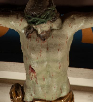 Zdjęcie nr 3: Krucyfiks zawieszony w łuku tęczowym kościoła. Do prostego krzyża przybita trzema gwoździami figura umarłego Chrystusa. Postać ukazana z szeroko i prosto rozłożonymi ramionami, z głową opadającą delikatnie w dół i z prawą nogą ugiętą w kolanie. Twarz Chrystusa jest podłużna, o zamkniętych oczach, długim i wąskim nosie, okolona jest krótką brodą rozdzieloną na końcu na dwa małe pukle i długimi włosami opadającymi na plecy i prawe ramię. Na głowie szeroka, zielona korona cierniowa. Ciało o delikatnie podkreślonej anatomii, smukłe. Perizonium zawiązane na prawym boku, odsłaniające prawe udo, z jednym końcem zwisającym wzdłuż prawego boku. Krzyż prosty, gładki, malowany na kolor ciemnobrązowy. Po jego lewej stronie, od tyłu nadlatujący anioł, nadnaturalnej wielkości, obejmujący rękami krzyż. Anioł ubrany w długą, złotą suknię, z rozłożonymi skrzydłami. Twarz anioła owalna, z otwartymi ustami, wzrokiem skierowanym na Chrystusa, nosem długim i spiczastym, silnie zarumienionymi policzkami, kręconymi włosami średniej długości. Nad głową Chrystusa na pionowej belce złota banderola z napisem „I.N./R.I.” Polichromia w odsłoniętych partiach ciała naturalistyczna, na figurze Chrystusa w odcieniach szarości, szaty i detale złocone. 
