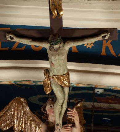 Zdjęcie nr 11: Krucyfiks zawieszony w łuku tęczowym kościoła. Do prostego krzyża przybita trzema gwoździami figura umarłego Chrystusa. Postać ukazana z szeroko i prosto rozłożonymi ramionami, z głową opadającą delikatnie w dół i z prawą nogą ugiętą w kolanie. Twarz Chrystusa jest podłużna, o zamkniętych oczach, długim i wąskim nosie, okolona jest krótką brodą rozdzieloną na końcu na dwa małe pukle i długimi włosami opadającymi na plecy i prawe ramię. Na głowie szeroka, zielona korona cierniowa. Ciało o delikatnie podkreślonej anatomii, smukłe. Perizonium zawiązane na prawym boku, odsłaniające prawe udo, z jednym końcem zwisającym wzdłuż prawego boku. Krzyż prosty, gładki, malowany na kolor ciemnobrązowy. Po jego lewej stronie, od tyłu nadlatujący anioł, nadnaturalnej wielkości, obejmujący rękami krzyż. Anioł ubrany w długą, złotą suknię, z rozłożonymi skrzydłami. Twarz anioła owalna, z otwartymi ustami, wzrokiem skierowanym na Chrystusa, nosem długim i spiczastym, silnie zarumienionymi policzkami, kręconymi włosami średniej długości. Nad głową Chrystusa na pionowej belce złota banderola z napisem „I.N./R.I.” Polichromia w odsłoniętych partiach ciała naturalistyczna, na figurze Chrystusa w odcieniach szarości, szaty i detale złocone. 
