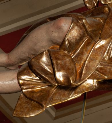 Zdjęcie nr 9: Krucyfiks zawieszony w łuku tęczowym kościoła. Do prostego krzyża przybita trzema gwoździami figura umarłego Chrystusa. Postać ukazana z szeroko i prosto rozłożonymi ramionami, z głową opadającą delikatnie w dół i z prawą nogą ugiętą w kolanie. Twarz Chrystusa jest podłużna, o zamkniętych oczach, długim i wąskim nosie, okolona jest krótką brodą rozdzieloną na końcu na dwa małe pukle i długimi włosami opadającymi na plecy i prawe ramię. Na głowie szeroka, zielona korona cierniowa. Ciało o delikatnie podkreślonej anatomii, smukłe. Perizonium zawiązane na prawym boku, odsłaniające prawe udo, z jednym końcem zwisającym wzdłuż prawego boku. Krzyż prosty, gładki, malowany na kolor ciemnobrązowy. Po jego lewej stronie, od tyłu nadlatujący anioł, nadnaturalnej wielkości, obejmujący rękami krzyż. Anioł ubrany w długą, złotą suknię, z rozłożonymi skrzydłami. Twarz anioła owalna, z otwartymi ustami, wzrokiem skierowanym na Chrystusa, nosem długim i spiczastym, silnie zarumienionymi policzkami, kręconymi włosami średniej długości. Nad głową Chrystusa na pionowej belce złota banderola z napisem „I.N./R.I.” Polichromia w odsłoniętych partiach ciała naturalistyczna, na figurze Chrystusa w odcieniach szarości, szaty i detale złocone. 
