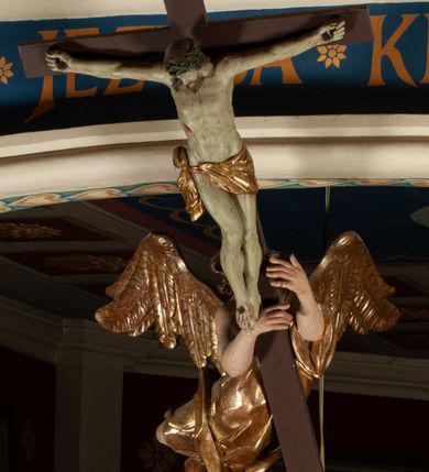 Zdjęcie nr 10: Krucyfiks zawieszony w łuku tęczowym kościoła. Do prostego krzyża przybita trzema gwoździami figura umarłego Chrystusa. Postać ukazana z szeroko i prosto rozłożonymi ramionami, z głową opadającą delikatnie w dół i z prawą nogą ugiętą w kolanie. Twarz Chrystusa jest podłużna, o zamkniętych oczach, długim i wąskim nosie, okolona jest krótką brodą rozdzieloną na końcu na dwa małe pukle i długimi włosami opadającymi na plecy i prawe ramię. Na głowie szeroka, zielona korona cierniowa. Ciało o delikatnie podkreślonej anatomii, smukłe. Perizonium zawiązane na prawym boku, odsłaniające prawe udo, z jednym końcem zwisającym wzdłuż prawego boku. Krzyż prosty, gładki, malowany na kolor ciemnobrązowy. Po jego lewej stronie, od tyłu nadlatujący anioł, nadnaturalnej wielkości, obejmujący rękami krzyż. Anioł ubrany w długą, złotą suknię, z rozłożonymi skrzydłami. Twarz anioła owalna, z otwartymi ustami, wzrokiem skierowanym na Chrystusa, nosem długim i spiczastym, silnie zarumienionymi policzkami, kręconymi włosami średniej długości. Nad głową Chrystusa na pionowej belce złota banderola z napisem „I.N./R.I.” Polichromia w odsłoniętych partiach ciała naturalistyczna, na figurze Chrystusa w odcieniach szarości, szaty i detale złocone. 
