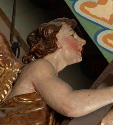 Zdjęcie nr 7: Krucyfiks zawieszony w łuku tęczowym kościoła. Do prostego krzyża przybita trzema gwoździami figura umarłego Chrystusa. Postać ukazana z szeroko i prosto rozłożonymi ramionami, z głową opadającą delikatnie w dół i z prawą nogą ugiętą w kolanie. Twarz Chrystusa jest podłużna, o zamkniętych oczach, długim i wąskim nosie, okolona jest krótką brodą rozdzieloną na końcu na dwa małe pukle i długimi włosami opadającymi na plecy i prawe ramię. Na głowie szeroka, zielona korona cierniowa. Ciało o delikatnie podkreślonej anatomii, smukłe. Perizonium zawiązane na prawym boku, odsłaniające prawe udo, z jednym końcem zwisającym wzdłuż prawego boku. Krzyż prosty, gładki, malowany na kolor ciemnobrązowy. Po jego lewej stronie, od tyłu nadlatujący anioł, nadnaturalnej wielkości, obejmujący rękami krzyż. Anioł ubrany w długą, złotą suknię, z rozłożonymi skrzydłami. Twarz anioła owalna, z otwartymi ustami, wzrokiem skierowanym na Chrystusa, nosem długim i spiczastym, silnie zarumienionymi policzkami, kręconymi włosami średniej długości. Nad głową Chrystusa na pionowej belce złota banderola z napisem „I.N./R.I.” Polichromia w odsłoniętych partiach ciała naturalistyczna, na figurze Chrystusa w odcieniach szarości, szaty i detale złocone. 
