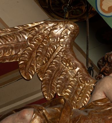 Zdjęcie nr 8: Krucyfiks zawieszony w łuku tęczowym kościoła. Do prostego krzyża przybita trzema gwoździami figura umarłego Chrystusa. Postać ukazana z szeroko i prosto rozłożonymi ramionami, z głową opadającą delikatnie w dół i z prawą nogą ugiętą w kolanie. Twarz Chrystusa jest podłużna, o zamkniętych oczach, długim i wąskim nosie, okolona jest krótką brodą rozdzieloną na końcu na dwa małe pukle i długimi włosami opadającymi na plecy i prawe ramię. Na głowie szeroka, zielona korona cierniowa. Ciało o delikatnie podkreślonej anatomii, smukłe. Perizonium zawiązane na prawym boku, odsłaniające prawe udo, z jednym końcem zwisającym wzdłuż prawego boku. Krzyż prosty, gładki, malowany na kolor ciemnobrązowy. Po jego lewej stronie, od tyłu nadlatujący anioł, nadnaturalnej wielkości, obejmujący rękami krzyż. Anioł ubrany w długą, złotą suknię, z rozłożonymi skrzydłami. Twarz anioła owalna, z otwartymi ustami, wzrokiem skierowanym na Chrystusa, nosem długim i spiczastym, silnie zarumienionymi policzkami, kręconymi włosami średniej długości. Nad głową Chrystusa na pionowej belce złota banderola z napisem „I.N./R.I.” Polichromia w odsłoniętych partiach ciała naturalistyczna, na figurze Chrystusa w odcieniach szarości, szaty i detale złocone. 
