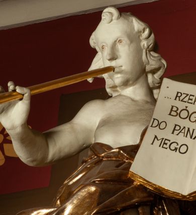 Zdjęcie nr 5: Rzeźba pełnoplastyczna, przedstawiająca anioła stojącego na obłoku z lewą nogą ugiętą w kolanie, który dmie w trąbę, a w lewej dłoni trzyma otwartą księgę z napisem „RZEKŁ / BÓG / DO PANA / MEGO / SIĄDŹ / PO PRAWICY / MOJEJ”. Twarz podłużna, okolona długimi, kręconymi włosami z wyraźnie zaznaczonym podbródkiem. Anioł owinięty jest bogato drapowaną tkaniną, tworzącą ostro załamujące się fałdy. Karnacja biała; szaty, skrzydła, trąbka i elementy księgi złocone.