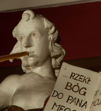 Zdjęcie nr 6: Rzeźba pełnoplastyczna, przedstawiająca anioła stojącego na obłoku z lewą nogą ugiętą w kolanie, który dmie w trąbę, a w lewej dłoni trzyma otwartą księgę z napisem „RZEKŁ / BÓG / DO PANA / MEGO / SIĄDŹ / PO PRAWICY / MOJEJ”. Twarz podłużna, okolona długimi, kręconymi włosami z wyraźnie zaznaczonym podbródkiem. Anioł owinięty jest bogato drapowaną tkaniną, tworzącą ostro załamujące się fałdy. Karnacja biała; szaty, skrzydła, trąbka i elementy księgi złocone.