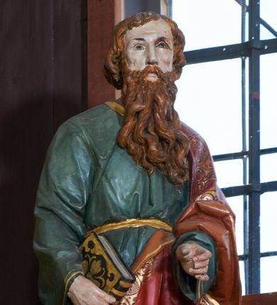 Zdjęcie nr 1: Rzeźba św. Pawła ustawiona na ośmiobocznej podstawie, wolnostojąca, pełnoplastyczna. Święty ukazany w całej postaci, stojący, z lewą nogą ugiętą w kolanie, z zamkniętą księgą w prawej dłoni oraz lewą ręką ugiętą w łokciu i wyciągniętą przed siebie. Twarz szeroka, z oczami skierowanymi w dal, okolona gęstą i długą brodą; włosy średniej długości, zasłaniające uszy. Ubrany w długą, zieloną suknię z długimi rękawami, ze złotą lamówką na brzegach, przewiązaną sznurem w okolicach talii. Na lewym ramieniu, plecach i z przodu sylwetki na wysokości kolan zarzucony długi, czerwony płaszcz z szeroką, dekoracyjną lamówką z geometrycznym wzorem; na nogach sandały. Polichromia w odsłoniętych partiach ciała naturalistyczna.

