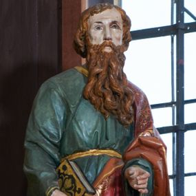 Zdjęcie nr 1: Rzeźba św. Pawła ustawiona na ośmiobocznej podstawie, wolnostojąca, pełnoplastyczna. Święty ukazany w całej postaci, stojący, z lewą nogą ugiętą w kolanie, z zamkniętą księgą w prawej dłoni oraz lewą ręką ugiętą w łokciu i wyciągniętą przed siebie. Twarz szeroka, z oczami skierowanymi w dal, okolona gęstą i długą brodą; włosy średniej długości, zasłaniające uszy. Ubrany w długą, zieloną suknię z długimi rękawami, ze złotą lamówką na brzegach, przewiązaną sznurem w okolicach talii. Na lewym ramieniu, plecach i z przodu sylwetki na wysokości kolan zarzucony długi, czerwony płaszcz z szeroką, dekoracyjną lamówką z geometrycznym wzorem; na nogach sandały. Polichromia w odsłoniętych partiach ciała naturalistyczna.


