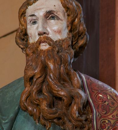 Zdjęcie nr 7: Rzeźba św. Pawła ustawiona na ośmiobocznej podstawie, wolnostojąca, pełnoplastyczna. Święty ukazany w całej postaci, stojący, z lewą nogą ugiętą w kolanie, z zamkniętą księgą w prawej dłoni oraz lewą ręką ugiętą w łokciu i wyciągniętą przed siebie. Twarz szeroka, z oczami skierowanymi w dal, okolona gęstą i długą brodą; włosy średniej długości, zasłaniające uszy. Ubrany w długą, zieloną suknię z długimi rękawami, ze złotą lamówką na brzegach, przewiązaną sznurem w okolicach talii. Na lewym ramieniu, plecach i z przodu sylwetki na wysokości kolan zarzucony długi, czerwony płaszcz z szeroką, dekoracyjną lamówką z geometrycznym wzorem; na nogach sandały. Polichromia w odsłoniętych partiach ciała naturalistyczna.

