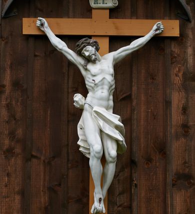 Zdjęcie nr 4: Pełnoplastyczna figura Chrystusa przybita do krzyża trzema gwoździami. Jezus o muskularnej budowie ciała z zaznaczonymi żebrami, wisi na krzyżu, ma wyprostowane ręce, które układają się na kształt litery „Y”,  prawa ręka ułożona jest pod nieco większym kątem niż lewa. Nogi ma lekko ugięte, głowę opartą na prawym ramieniu, oczy zamknięte. Ciemnobrązowe włosy spływają na prawe ramię, twarz okala krótki zarost. Na głowie ma założoną koronę cierniową w kolorze włosów i zarostu. Po czole oraz z ran na rękach, nogach i prawym boku spływają ciemnoczerwone strużki krwi. Chrystus odziany jest w białe perizonium, które zawieszone jest na podwójnym sznurze. Karnacja ciała biała, krzyż jasnobrązowy, na górze tabliczka z napisem „I.N. / R.I”. Nad krzyżem wisi półkolisty, blaszany, brązowy daszek.