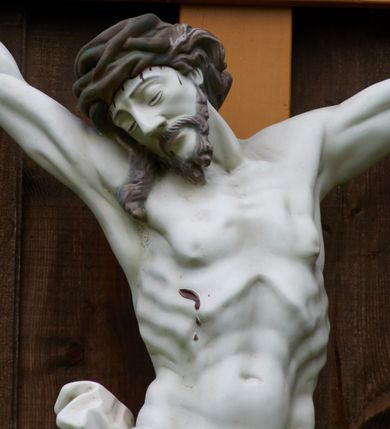 Zdjęcie nr 5: Pełnoplastyczna figura Chrystusa przybita do krzyża trzema gwoździami. Jezus o muskularnej budowie ciała z zaznaczonymi żebrami, wisi na krzyżu, ma wyprostowane ręce, które układają się na kształt litery „Y”,  prawa ręka ułożona jest pod nieco większym kątem niż lewa. Nogi ma lekko ugięte, głowę opartą na prawym ramieniu, oczy zamknięte. Ciemnobrązowe włosy spływają na prawe ramię, twarz okala krótki zarost. Na głowie ma założoną koronę cierniową w kolorze włosów i zarostu. Po czole oraz z ran na rękach, nogach i prawym boku spływają ciemnoczerwone strużki krwi. Chrystus odziany jest w białe perizonium, które zawieszone jest na podwójnym sznurze. Karnacja ciała biała, krzyż jasnobrązowy, na górze tabliczka z napisem „I.N. / R.I”. Nad krzyżem wisi półkolisty, blaszany, brązowy daszek.
