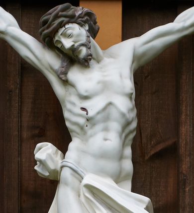 Zdjęcie nr 1: Pełnoplastyczna figura Chrystusa przybita do krzyża trzema gwoździami. Jezus o muskularnej budowie ciała z zaznaczonymi żebrami, wisi na krzyżu, ma wyprostowane ręce, które układają się na kształt litery „Y”,  prawa ręka ułożona jest pod nieco większym kątem niż lewa. Nogi ma lekko ugięte, głowę opartą na prawym ramieniu, oczy zamknięte. Ciemnobrązowe włosy spływają na prawe ramię, twarz okala krótki zarost. Na głowie ma założoną koronę cierniową w kolorze włosów i zarostu. Po czole oraz z ran na rękach, nogach i prawym boku spływają ciemnoczerwone strużki krwi. Chrystus odziany jest w białe perizonium, które zawieszone jest na podwójnym sznurze. Karnacja ciała biała, krzyż jasnobrązowy, na górze tabliczka z napisem „I.N. / R.I”. Nad krzyżem wisi półkolisty, blaszany, brązowy daszek.