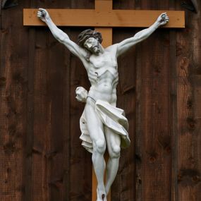 Zdjęcie nr 1: Pełnoplastyczna figura Chrystusa przybita do krzyża trzema gwoździami. Jezus o muskularnej budowie ciała z zaznaczonymi żebrami, wisi na krzyżu, ma wyprostowane ręce, które układają się na kształt litery „Y”,  prawa ręka ułożona jest pod nieco większym kątem niż lewa. Nogi ma lekko ugięte, głowę opartą na prawym ramieniu, oczy zamknięte. Ciemnobrązowe włosy spływają na prawe ramię, twarz okala krótki zarost. Na głowie ma założoną koronę cierniową w kolorze włosów i zarostu. Po czole oraz z ran na rękach, nogach i prawym boku spływają ciemnoczerwone strużki krwi. Chrystus odziany jest w białe perizonium, które zawieszone jest na podwójnym sznurze. Karnacja ciała biała, krzyż jasnobrązowy, na górze tabliczka z napisem „I.N. / R.I”. Nad krzyżem wisi półkolisty, blaszany, brązowy daszek.