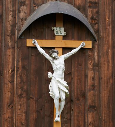 Zdjęcie nr 3: Pełnoplastyczna figura Chrystusa przybita do krzyża trzema gwoździami. Jezus o muskularnej budowie ciała z zaznaczonymi żebrami, wisi na krzyżu, ma wyprostowane ręce, które układają się na kształt litery „Y”,  prawa ręka ułożona jest pod nieco większym kątem niż lewa. Nogi ma lekko ugięte, głowę opartą na prawym ramieniu, oczy zamknięte. Ciemnobrązowe włosy spływają na prawe ramię, twarz okala krótki zarost. Na głowie ma założoną koronę cierniową w kolorze włosów i zarostu. Po czole oraz z ran na rękach, nogach i prawym boku spływają ciemnoczerwone strużki krwi. Chrystus odziany jest w białe perizonium, które zawieszone jest na podwójnym sznurze. Karnacja ciała biała, krzyż jasnobrązowy, na górze tabliczka z napisem „I.N. / R.I”. Nad krzyżem wisi półkolisty, blaszany, brązowy daszek.