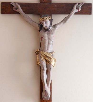Zdjęcie nr 1: Rzeźba Chrystusa przybita do krzyża trzema gwoździami. Chrystus ukazany w typie Cristo morte, o delikatnie esowato wygiętej sylwetce, w lekkim zwisie, z głową nieznacznie pochyloną na prawą stronę. Chrystus ma masywne dłonie i stopy, wyraźnie podkreśloną linię żeber, sylwetkę szczupłą. Jego twarz jest podłużna, okolona krótką brodą, ciemnobrązowe włosy spływają na prawe ramię. Na głowie ma założoną koronę cierniową. Jego biodra oplata złocone perizonium zawieszone na sznurze. Z dłoni, stóp i boku Chrystusa spływają strużki krwi. Polichromia ciała naturalistyczna, detale korona cierniowa i perizonium złocone. Krzyż prosty, gładki, malowany w kolorze drewna. 
