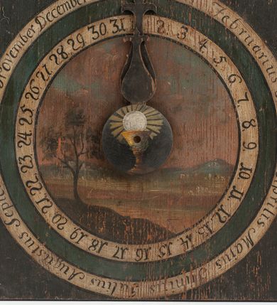 Zdjęcie nr 3: Zegar w kształcie stojącego prostokąta. W części centralnej okrągła tarcza z pejzażem oraz kołem pośrodku, ozdobionym kielichem z hostią, od którego odchodzą dwie fantazyjnie wycięte wskazówki. Wokół tarczy z pejzażem dwa okręgi, w pierwszym liczby od 1 do 31, w drugim, zewnętrznym – łacińskie nazwy miesięcy. W górnej części kompozycji biała banderola z napisem: „Renovatio Sanctissimi Sacramenti”. Poniżej, wpisany w koło na czerwonym tle monogram Chrystusa. Tło czarne.

