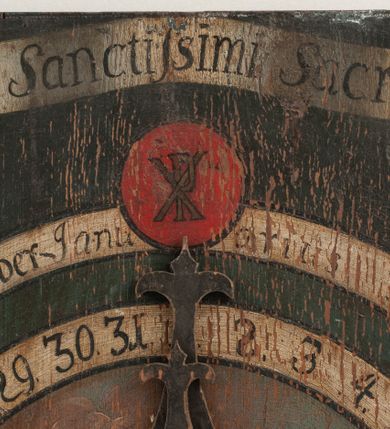 Zdjęcie nr 4: Zegar w kształcie stojącego prostokąta. W części centralnej okrągła tarcza z pejzażem oraz kołem pośrodku, ozdobionym kielichem z hostią, od którego odchodzą dwie fantazyjnie wycięte wskazówki. Wokół tarczy z pejzażem dwa okręgi, w pierwszym liczby od 1 do 31, w drugim, zewnętrznym – łacińskie nazwy miesięcy. W górnej części kompozycji biała banderola z napisem: „Renovatio Sanctissimi Sacramenti”. Poniżej, wpisany w koło na czerwonym tle monogram Chrystusa. Tło czarne.

