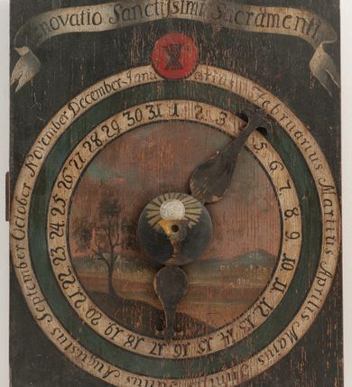 Zdjęcie nr 2: Zegar w kształcie stojącego prostokąta. W części centralnej okrągła tarcza z pejzażem oraz kołem pośrodku, ozdobionym kielichem z hostią, od którego odchodzą dwie fantazyjnie wycięte wskazówki. Wokół tarczy z pejzażem dwa okręgi, w pierwszym liczby od 1 do 31, w drugim, zewnętrznym – łacińskie nazwy miesięcy. W górnej części kompozycji biała banderola z napisem: „Renovatio Sanctissimi Sacramenti”. Poniżej, wpisany w koło na czerwonym tle monogram Chrystusa. Tło czarne.

