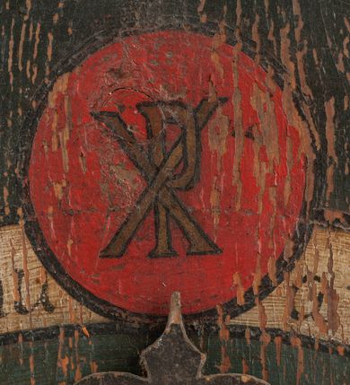 Zdjęcie nr 5: Zegar w kształcie stojącego prostokąta. W części centralnej okrągła tarcza z pejzażem oraz kołem pośrodku, ozdobionym kielichem z hostią, od którego odchodzą dwie fantazyjnie wycięte wskazówki. Wokół tarczy z pejzażem dwa okręgi, w pierwszym liczby od 1 do 31, w drugim, zewnętrznym – łacińskie nazwy miesięcy. W górnej części kompozycji biała banderola z napisem: „Renovatio Sanctissimi Sacramenti”. Poniżej, wpisany w koło na czerwonym tle monogram Chrystusa. Tło czarne.

