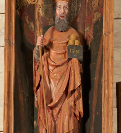 Zdjęcie nr 2: Rzeźba pełnoplastyczna, przyścienna, ścięta z tyłu; całopostaciowe przedstawienie stojącego na niewysokim postumencie św. Mikołaja. Święty ukazany frontalnie. Twarz pociągła, o wyrazistych rysach i długim, brązowym zaroście, włosy sięgające szyi. Święty w stroju pontyfikalnym: ubrany w długą, białą albę oraz jasnoczerwoną dalmatykę lub tunicelę i ornat sięgający do połowy łydek. Na głowie infuła biskupia. W lewej ręce prostopadłościenna skrzyneczka z napisem: IHS i trzema złotymi kulami, w prawej pastorał. Figura umieszczona w prostopadłościennej szafie oltarzowej o skośnych ścianach bocznych zdobionych figuralną polichromią. Po lewej stronie św. Wawrzyniec, po prawej św. Szczepan, na wysokości głowy świętego polichromowana, jasna aureola.