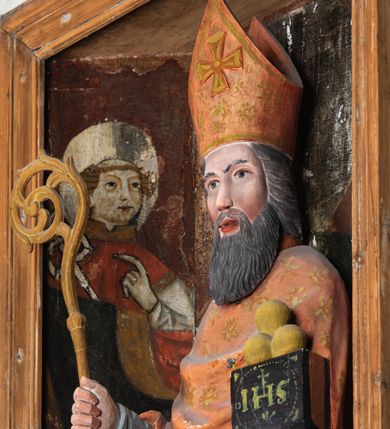 Zdjęcie nr 5: Rzeźba pełnoplastyczna, przyścienna, ścięta z tyłu; całopostaciowe przedstawienie stojącego na niewysokim postumencie św. Mikołaja. Święty ukazany frontalnie. Twarz pociągła, o wyrazistych rysach i długim, brązowym zaroście, włosy sięgające szyi. Święty w stroju pontyfikalnym: ubrany w długą, białą albę oraz jasnoczerwoną dalmatykę lub tunicelę i ornat sięgający do połowy łydek. Na głowie infuła biskupia. W lewej ręce prostopadłościenna skrzyneczka z napisem: IHS i trzema złotymi kulami, w prawej pastorał. Figura umieszczona w prostopadłościennej szafie oltarzowej o skośnych ścianach bocznych zdobionych figuralną polichromią. Po lewej stronie św. Wawrzyniec, po prawej św. Szczepan, na wysokości głowy świętego polichromowana, jasna aureola.