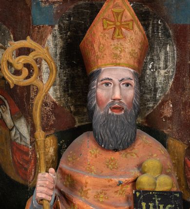 Zdjęcie nr 4: Rzeźba pełnoplastyczna, przyścienna, ścięta z tyłu; całopostaciowe przedstawienie stojącego na niewysokim postumencie św. Mikołaja. Święty ukazany frontalnie. Twarz pociągła, o wyrazistych rysach i długim, brązowym zaroście, włosy sięgające szyi. Święty w stroju pontyfikalnym: ubrany w długą, białą albę oraz jasnoczerwoną dalmatykę lub tunicelę i ornat sięgający do połowy łydek. Na głowie infuła biskupia. W lewej ręce prostopadłościenna skrzyneczka z napisem: IHS i trzema złotymi kulami, w prawej pastorał. Figura umieszczona w prostopadłościennej szafie oltarzowej o skośnych ścianach bocznych zdobionych figuralną polichromią. Po lewej stronie św. Wawrzyniec, po prawej św. Szczepan, na wysokości głowy świętego polichromowana, jasna aureola.