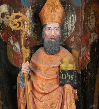 Zdjęcie nr 3: Rzeźba pełnoplastyczna, przyścienna, ścięta z tyłu; całopostaciowe przedstawienie stojącego na niewysokim postumencie św. Mikołaja. Święty ukazany frontalnie. Twarz pociągła, o wyrazistych rysach i długim, brązowym zaroście, włosy sięgające szyi. Święty w stroju pontyfikalnym: ubrany w długą, białą albę oraz jasnoczerwoną dalmatykę lub tunicelę i ornat sięgający do połowy łydek. Na głowie infuła biskupia. W lewej ręce prostopadłościenna skrzyneczka z napisem: IHS i trzema złotymi kulami, w prawej pastorał. Figura umieszczona w prostopadłościennej szafie oltarzowej o skośnych ścianach bocznych zdobionych figuralną polichromią. Po lewej stronie św. Wawrzyniec, po prawej św. Szczepan, na wysokości głowy świętego polichromowana, jasna aureola.