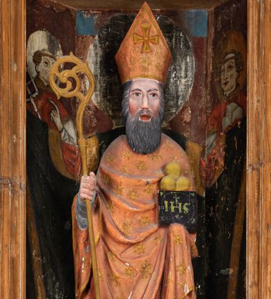 Zdjęcie nr 1: Rzeźba pełnoplastyczna, przyścienna, ścięta z tyłu; całopostaciowe przedstawienie stojącego na niewysokim postumencie św. Mikołaja. Święty ukazany frontalnie. Twarz pociągła, o wyrazistych rysach i długim, brązowym zaroście, włosy sięgające szyi. Święty w stroju pontyfikalnym: ubrany w długą, białą albę oraz jasnoczerwoną dalmatykę lub tunicelę i ornat sięgający do połowy łydek. Na głowie infuła biskupia. W lewej ręce prostopadłościenna skrzyneczka z napisem: IHS i trzema złotymi kulami, w prawej pastorał. Figura umieszczona w prostopadłościennej szafie oltarzowej o skośnych ścianach bocznych zdobionych figuralną polichromią. Po lewej stronie św. Wawrzyniec, po prawej św. Szczepan, na wysokości głowy świętego polichromowana, jasna aureola.