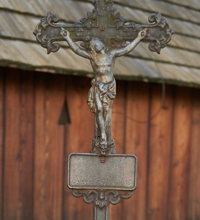 Zdjęcie nr 1: Żeliwny krucyfiks na kamiennym, graniastosłupowym postumencie zakończonym półkuliście. Metalowa, półplastyczna figura Ukrzyżowanego ubranego w krótkie perizonium związane po lewej stronie. Ramiona tworzą kształt litery U; głowa przechylona w prawo. Nogi przebite jednym gwoździem. Zakończenia i przecięcie ramion krzyża zdobione motywami roślinnymi. W górnej części belki pionowej titulus z literami: I.N.R.I.;  poniżej stóp Chrystusa tabliczka w kształcie leżącego prostokąta, z  wolutowo - roślinnym podwieszeniem (fartuchem). Podstawa krzyża w formie symetrycznego ornamentu roślinnego z dwoma dużymi kwiatami, na których stoi para aniołków ustawionych antytetycznie, ubranych w krótkie tuniki, ze złożonymi rękami.