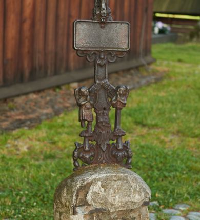 Zdjęcie nr 2: Żeliwny krucyfiks na kamiennym, graniastosłupowym postumencie zakończonym półkuliście. Metalowa, półplastyczna figura Ukrzyżowanego ubranego w krótkie perizonium związane po lewej stronie. Ramiona tworzą kształt litery U; głowa przechylona w prawo. Nogi przebite jednym gwoździem. Zakończenia i przecięcie ramion krzyża zdobione motywami roślinnymi. W górnej części belki pionowej titulus z literami: I.N.R.I.;  poniżej stóp Chrystusa tabliczka w kształcie leżącego prostokąta, z  wolutowo - roślinnym podwieszeniem (fartuchem). Podstawa krzyża w formie symetrycznego ornamentu roślinnego z dwoma dużymi kwiatami, na których stoi para aniołków ustawionych antytetycznie, ubranych w krótkie tuniki, ze złożonymi rękami.