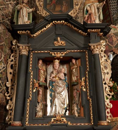 Zdjęcie nr 6: Obraz w kształcie stojącego ośmioboku z przedstawieniem Zwiastowania NMP, we wnętrzu z oknem w tle. W prawej dolnej części obrazu klęcząca na klęczniku Maria w ujęciu trzy czwarte, zwracająca głowę w stronę unoszącego się tuż nad posadzką Archanioła Gabriela. Archanioł znajduje się w lewej części obrazu, ukazany z profilu, zwraca się w stronę Marii, prawą rękę unosi do góry, w lewej trzyma białą lilię. Scena ukazana we wnętrzu. W górnej partii obrazu świetlisty obłok z gołębicą Ducha Świętego. Kolorystyka utrzymana w tonacji ciepłej, z przewagą brązów, ugrów, bieli, żółcieni i czerwieni.