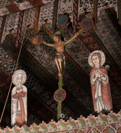Zdjęcie nr 8: Pośrodku belki tęczowej krzyż widlasty z figurą martwego Chrystusa Ukrzyżowanego, flankowany przez malowane na desce postacie Matki Boskiej i św. Jana Ewangelisty. Obrazy w kształcie wyciętego w desce konturu postaci. Krzyż w formie drzewa życia, z pojedynczymi wypustkami, na zakończeniach ramion medaliony z przedstawieniami symboli Ewangelistów z banderolami, górny medalion pusty; pod nim ukośny titulus w formie banderoli z napisem: INRI. Figura Chrystusa w lekkim zwisie, o szeroko rozłożonych ramionach, głowa opadająca; nogi silnie zgięte w kolanach, podkulone; stopy przebite jednym gwoździem, założone prawa na lewą. Ciało o niewłaściwych proporcjach anatomicznych, z silnie podkreśloną klatką piersiową oraz zaznaczonymi śladami męki. Twarz pociągła, o delikatnie rzeźbionych rysach, okolona zarostem. Włosy długie, falowane, opadają na plecy, na głowie korona cierniowa. Perizonium długie, sięgające kolan, ściśle opina biodra, z dwoma zwisami tkaniny po bokach. Polichromia naturalistyczna.