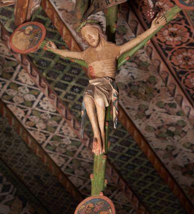 Zdjęcie nr 7: Pośrodku belki tęczowej krzyż widlasty z figurą martwego Chrystusa Ukrzyżowanego, flankowany przez malowane na desce postacie Matki Boskiej i św. Jana Ewangelisty. Obrazy w kształcie wyciętego w desce konturu postaci. Krzyż w formie drzewa życia, z pojedynczymi wypustkami, na zakończeniach ramion medaliony z przedstawieniami symboli Ewangelistów z banderolami, górny medalion pusty; pod nim ukośny titulus w formie banderoli z napisem: INRI. Figura Chrystusa w lekkim zwisie, o szeroko rozłożonych ramionach, głowa opadająca; nogi silnie zgięte w kolanach, podkulone; stopy przebite jednym gwoździem, założone prawa na lewą. Ciało o niewłaściwych proporcjach anatomicznych, z silnie podkreśloną klatką piersiową oraz zaznaczonymi śladami męki. Twarz pociągła, o delikatnie rzeźbionych rysach, okolona zarostem. Włosy długie, falowane, opadają na plecy, na głowie korona cierniowa. Perizonium długie, sięgające kolan, ściśle opina biodra, z dwoma zwisami tkaniny po bokach. Polichromia naturalistyczna.