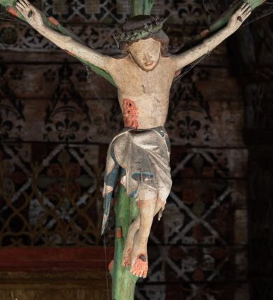 Zdjęcie nr 3: Pośrodku belki tęczowej krzyż widlasty z figurą martwego Chrystusa Ukrzyżowanego, flankowany przez malowane na desce postacie Matki Boskiej i św. Jana Ewangelisty. Obrazy w kształcie wyciętego w desce konturu postaci. Krzyż w formie drzewa życia, z pojedynczymi wypustkami, na zakończeniach ramion medaliony z przedstawieniami symboli Ewangelistów z banderolami, górny medalion pusty; pod nim ukośny titulus w formie banderoli z napisem: INRI. Figura Chrystusa w lekkim zwisie, o szeroko rozłożonych ramionach, głowa opadająca; nogi silnie zgięte w kolanach, podkulone; stopy przebite jednym gwoździem, założone prawa na lewą. Ciało o niewłaściwych proporcjach anatomicznych, z silnie podkreśloną klatką piersiową oraz zaznaczonymi śladami męki. Twarz pociągła, o delikatnie rzeźbionych rysach, okolona zarostem. Włosy długie, falowane, opadają na plecy, na głowie korona cierniowa. Perizonium długie, sięgające kolan, ściśle opina biodra, z dwoma zwisami tkaniny po bokach. Polichromia naturalistyczna.