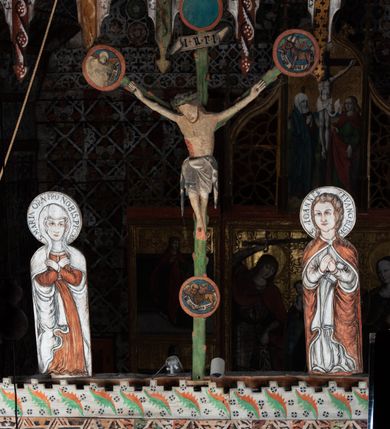 Zdjęcie nr 2: Pośrodku belki tęczowej krzyż widlasty z figurą martwego Chrystusa Ukrzyżowanego, flankowany przez malowane na desce postacie Matki Boskiej i św. Jana Ewangelisty. Obrazy w kształcie wyciętego w desce konturu postaci. Krzyż w formie drzewa życia, z pojedynczymi wypustkami, na zakończeniach ramion medaliony z przedstawieniami symboli Ewangelistów z banderolami, górny medalion pusty; pod nim ukośny titulus w formie banderoli z napisem: INRI. Figura Chrystusa w lekkim zwisie, o szeroko rozłożonych ramionach, głowa opadająca; nogi silnie zgięte w kolanach, podkulone; stopy przebite jednym gwoździem, założone prawa na lewą. Ciało o niewłaściwych proporcjach anatomicznych, z silnie podkreśloną klatką piersiową oraz zaznaczonymi śladami męki. Twarz pociągła, o delikatnie rzeźbionych rysach, okolona zarostem. Włosy długie, falowane, opadają na plecy, na głowie korona cierniowa. Perizonium długie, sięgające kolan, ściśle opina biodra, z dwoma zwisami tkaniny po bokach. Polichromia naturalistyczna.