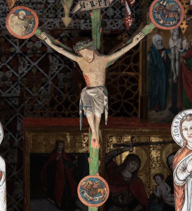 Zdjęcie nr 1: Pośrodku belki tęczowej krzyż widlasty z figurą martwego Chrystusa Ukrzyżowanego, flankowany przez malowane na desce postacie Matki Boskiej i św. Jana Ewangelisty. Obrazy w kształcie wyciętego w desce konturu postaci. Krzyż w formie drzewa życia, z pojedynczymi wypustkami, na zakończeniach ramion medaliony z przedstawieniami symboli Ewangelistów z banderolami, górny medalion pusty; pod nim ukośny titulus w formie banderoli z napisem: INRI. Figura Chrystusa w lekkim zwisie, o szeroko rozłożonych ramionach, głowa opadająca; nogi silnie zgięte w kolanach, podkulone; stopy przebite jednym gwoździem, założone prawa na lewą. Ciało o niewłaściwych proporcjach anatomicznych, z silnie podkreśloną klatką piersiową oraz zaznaczonymi śladami męki. Twarz pociągła, o delikatnie rzeźbionych rysach, okolona zarostem. Włosy długie, falowane, opadają na plecy, na głowie korona cierniowa. Perizonium długie, sięgające kolan, ściśle opina biodra, z dwoma zwisami tkaniny po bokach. Polichromia naturalistyczna.