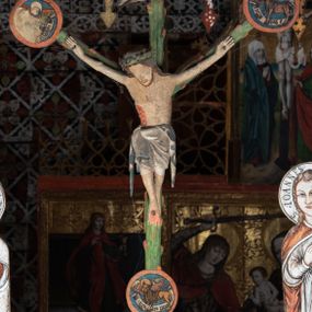 Zdjęcie nr 1: Pośrodku belki tęczowej krzyż widlasty z figurą martwego Chrystusa Ukrzyżowanego, flankowany przez malowane na desce postacie Matki Boskiej i św. Jana Ewangelisty. Obrazy w kształcie wyciętego w desce konturu postaci. Krzyż w formie drzewa życia, z pojedynczymi wypustkami, na zakończeniach ramion medaliony z przedstawieniami symboli Ewangelistów z banderolami, górny medalion pusty; pod nim ukośny titulus w formie banderoli z napisem: INRI. Figura Chrystusa w lekkim zwisie, o szeroko rozłożonych ramionach, głowa opadająca; nogi silnie zgięte w kolanach, podkulone; stopy przebite jednym gwoździem, założone prawa na lewą. Ciało o niewłaściwych proporcjach anatomicznych, z silnie podkreśloną klatką piersiową oraz zaznaczonymi śladami męki. Twarz pociągła, o delikatnie rzeźbionych rysach, okolona zarostem. Włosy długie, falowane, opadają na plecy, na głowie korona cierniowa. Perizonium długie, sięgające kolan, ściśle opina biodra, z dwoma zwisami tkaniny po bokach. Polichromia naturalistyczna.