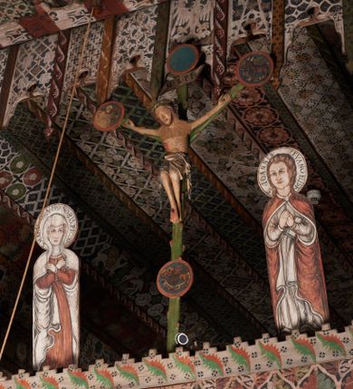 Zdjęcie nr 3: W centrum krzyż widlasty z figurą martwego Chrystusa Ukrzyżowanego; flankowany przez malowane na desce postacie Matki Boskiej i św. Jana. Obrazy w kształcie wyciętego obrysu postaci. Obraz z Matką Boską po prawej stronie krzyża. Maria zwrócona en face, z rękoma skrzyżowanymi na piersi i głową skierowaną w stronę krzyża. Twarz owalna,, o młodzieńczych rysach. Ubrana w czerwoną suknię z długimi, wąskimi rękawami ze złotą lamówką oraz biały płaszcz narzucony na głowę i ramiona, opadający falami po bokach. Wokół głowy nimb z napisem: S. MARIA ORA PRO NOBIS PECCATOR[IBUS] Obraz z przedstawieniem św. Jana po lewej stronie krzyża. Święty zwrócony en face, z dłońmi złożonymi na piersi, lewą podtrzymuje zamkniętą księgę. Twarz owalna, o pełnych policzkach i wyraźnych, młodzieńczych rysach. Włosy długie poskręcane w pukle i opadające na ramiona. Ubrany w białą tunikę o wąskich długich rękawach, drapowaną w równoległe fałdy oraz czerwony płaszcz zapięty przy szyi fibulą, opadający kaskadami na ramiona i boki. Wokół głowy nimb z napisem: S. IOANNES EVANGELISTA. Malowane płaską plamą barwną z silnym konturem.