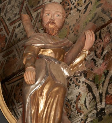 Zdjęcie nr 4: Rzeźba pełnoplastyczna, drążona, ustawiona na prostopadłościennym cokole. Święty ukazany frontalnie, w wykroku, przechylony do tyłu, opierając się o stojący na nim krzyż w kształcie litery X; lewą ręką podtrzymuje belkę, prawa pozostaje opuszczona wzdłuż ciała. Święty ubrany w złotą, drapowaną tunikę z długim rękawem oraz srebrny płaszcz przerzucony przez pierś i zawinięty na biodrze.