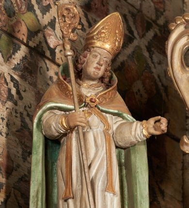 Zdjęcie nr 4: Rzeźba pełnoplastyczna, ścięta z tyłu, drążona. Święty ukazany en face, z głową przechyloną na lewe ramię. W prawej ręce trzyma pastorał, lewa wyciągnięta przed siebie. Twarz pociągła, oczy głęboko osadzone, nos prosty. Włosy  kręcone, opadające na ramiona. Ubrany w białą albę, drapowaną w sztywne, rurkowe fałdy, i skrzyżowaną na piersi stułę, przepasane w talii; oraz złoconą, sztywną kapę liturgiczną, podbitą zieloną podszewką, spiętą na piersi fibulą. Na głowie infuła biskupia o rzeźbionej dekoracji. Przy lewej nodze świętego półpostać Piotrowina, ukazanego z profilu ze złożonymi w geście modlitwy dłońmi. Polichromia naturalistyczna, atrybuty złocone.