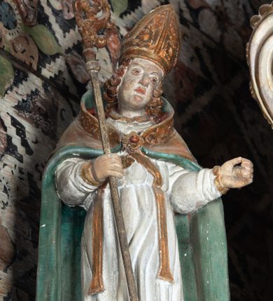 Zdjęcie nr 1: Rzeźba pełnoplastyczna, ścięta z tyłu, drążona. Święty ukazany en face, z głową przechyloną na lewe ramię. W prawej ręce trzyma pastorał, lewa wyciągnięta przed siebie. Twarz pociągła, oczy głęboko osadzone, nos prosty. Włosy  kręcone, opadające na ramiona. Ubrany w białą albę, drapowaną w sztywne, rurkowe fałdy, i skrzyżowaną na piersi stułę, przepasane w talii; oraz złoconą, sztywną kapę liturgiczną, podbitą zieloną podszewką, spiętą na piersi fibulą. Na głowie infuła biskupia o rzeźbionej dekoracji. Przy lewej nodze świętego półpostać Piotrowina, ukazanego z profilu ze złożonymi w geście modlitwy dłońmi. Polichromia naturalistyczna, atrybuty złocone.