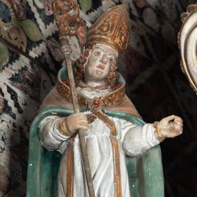 Zdjęcie nr 1: Rzeźba pełnoplastyczna, ścięta z tyłu, drążona. Święty ukazany en face, z głową przechyloną na lewe ramię. W prawej ręce trzyma pastorał, lewa wyciągnięta przed siebie. Twarz pociągła, oczy głęboko osadzone, nos prosty. Włosy  kręcone, opadające na ramiona. Ubrany w białą albę, drapowaną w sztywne, rurkowe fałdy, i skrzyżowaną na piersi stułę, przepasane w talii; oraz złoconą, sztywną kapę liturgiczną, podbitą zieloną podszewką, spiętą na piersi fibulą. Na głowie infuła biskupia o rzeźbionej dekoracji. Przy lewej nodze świętego półpostać Piotrowina, ukazanego z profilu ze złożonymi w geście modlitwy dłońmi. Polichromia naturalistyczna, atrybuty złocone.