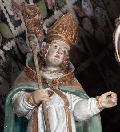 Zdjęcie nr 2: Rzeźba pełnoplastyczna, ścięta z tyłu, drążona. Święty ukazany en face, z głową przechyloną na lewe ramię. W prawej ręce trzyma pastorał, lewa wyciągnięta przed siebie. Twarz pociągła, oczy głęboko osadzone, nos prosty. Włosy  kręcone, opadające na ramiona. Ubrany w białą albę, drapowaną w sztywne, rurkowe fałdy, i skrzyżowaną na piersi stułę, przepasane w talii; oraz złoconą, sztywną kapę liturgiczną, podbitą zieloną podszewką, spiętą na piersi fibulą. Na głowie infuła biskupia o rzeźbionej dekoracji. Przy lewej nodze świętego półpostać Piotrowina, ukazanego z profilu ze złożonymi w geście modlitwy dłońmi. Polichromia naturalistyczna, atrybuty złocone.